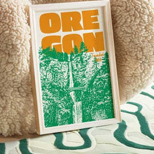 May include: Framed art print with the word "OREGON" in orange block letters above a green and white illustration of a waterfall and trees. The print is in a white frame and rests on a patterned surface.