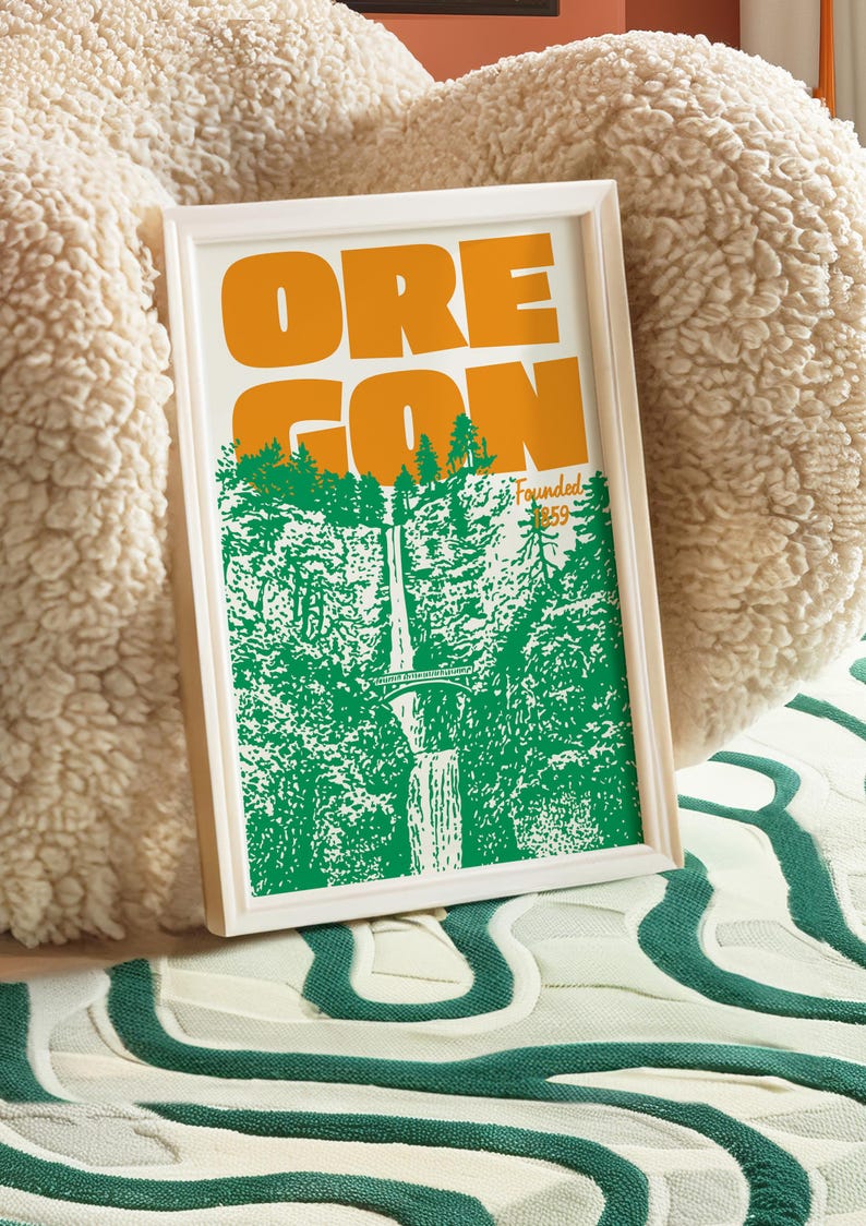 May include: Framed art print with the word "OREGON" in orange block letters above a green and white illustration of a waterfall and trees. The print is in a white frame and rests on a patterned surface.