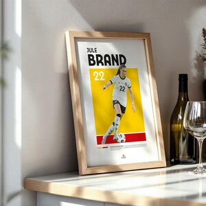 Może przedstawiać: Oprawiony plakat z piłkarką Jule Brand w białej koszulce z numerem 22. Plakat ma ż&oacute;łto-czerwone tło. Butelka wina i kieliszek znajdują się w tle.