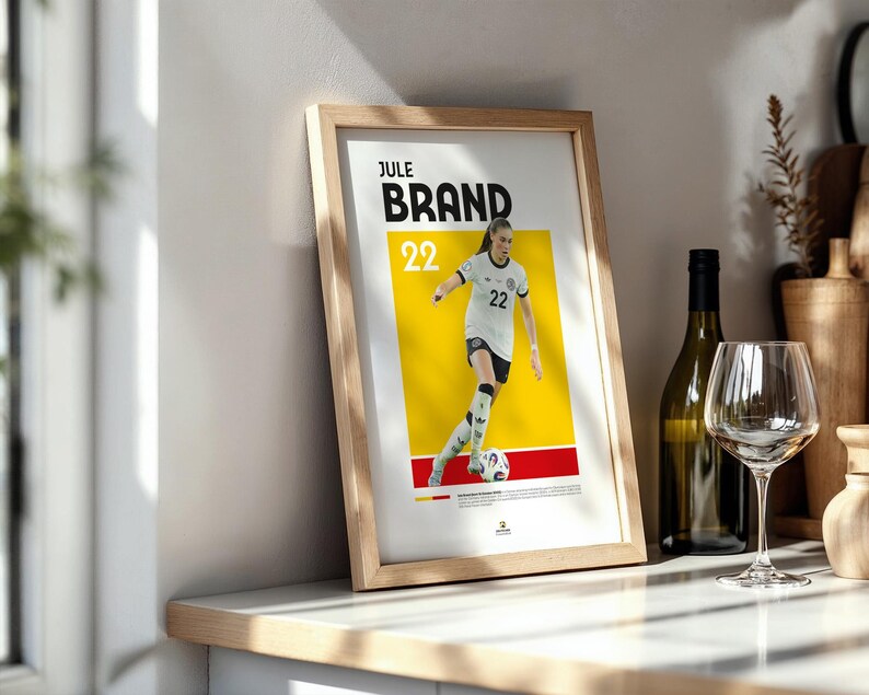 Może przedstawiać: Oprawiony plakat z piłkarką Jule Brand w białej koszulce z numerem 22. Plakat ma ż&oacute;łto-czerwone tło. Butelka wina i kieliszek znajdują się w tle.