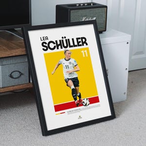 May include: Framed poster of Lea Sch&uuml;ller, a footballer, in action. The poster has a yellow and red background with the player's name and number 11 in bold text. The player is wearing a white and black uniform.