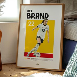 Może przedstawiać: Oprawiony plakat z Jule Brand, piłkarką nożną, w biało-czarnym stroju z numerem 22. Plakat ma ż&oacute;łto-czerwone tło. Plakat jest w drewnianej ramie.
