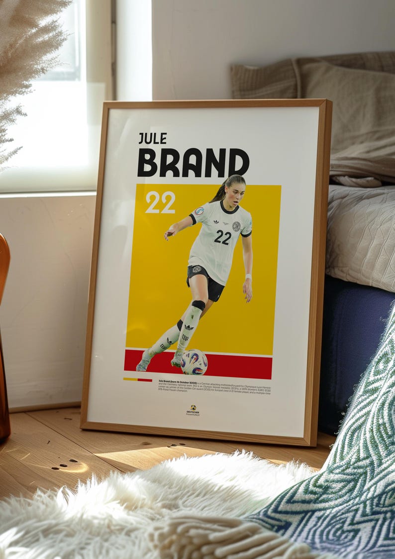 Może przedstawiać: Oprawiony plakat z Jule Brand, piłkarką nożną, w biało-czarnym stroju z numerem 22. Plakat ma ż&oacute;łto-czerwone tło. Plakat jest w drewnianej ramie.