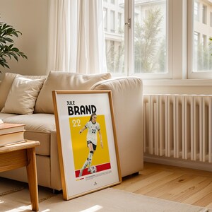 Może przedstawiać: Oprawiony plakat z piłkarką Jule Brand w biało-czarnym stroju na ż&oacute;łto-czerwonym tle. Plakat zawiera imię "BRAND" i numer "22". Plakat znajduje się w salonie z beżową sofą i drewnianą podłogą.