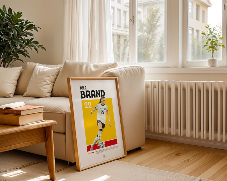 Może przedstawiać: Oprawiony plakat z piłkarką Jule Brand w biało-czarnym stroju na ż&oacute;łto-czerwonym tle. Plakat zawiera imię "BRAND" i numer "22". Plakat znajduje się w salonie z beżową sofą i drewnianą podłogą.