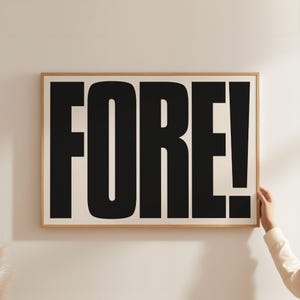May include: A framed art print with the word "FORE!" in bold, black letters against a cream background. The wooden frame surrounds the print, and a person's hand is visible, holding the frame.