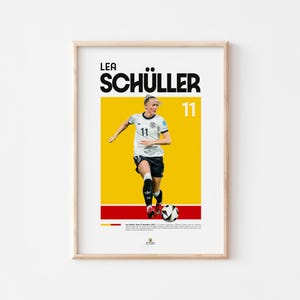 May include: Framed poster of a soccer player in a white and black uniform, with the name "LEA SCH&Uuml;LLER" in bold black letters. The number "11" is displayed. The background is yellow with red and black stripes at the bottom.