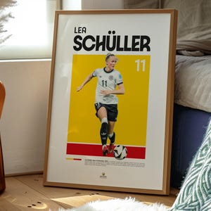 May include: Wooden framed poster of Lea Sch&uuml;ller, number 11, in a football uniform. The poster has a yellow background with the player's name in large black letters. The image is set in a room with a wooden floor and a white rug.