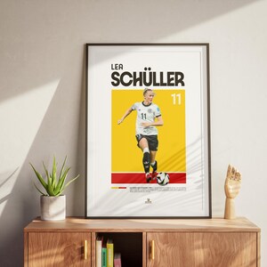 May include: Framed poster of footballer Lea Sch&uuml;ller in action, set against a yellow and red background. The poster displays her name and the number 11. The image is displayed on a neutral wall, with a wooden cabinet and plant in the foreground.