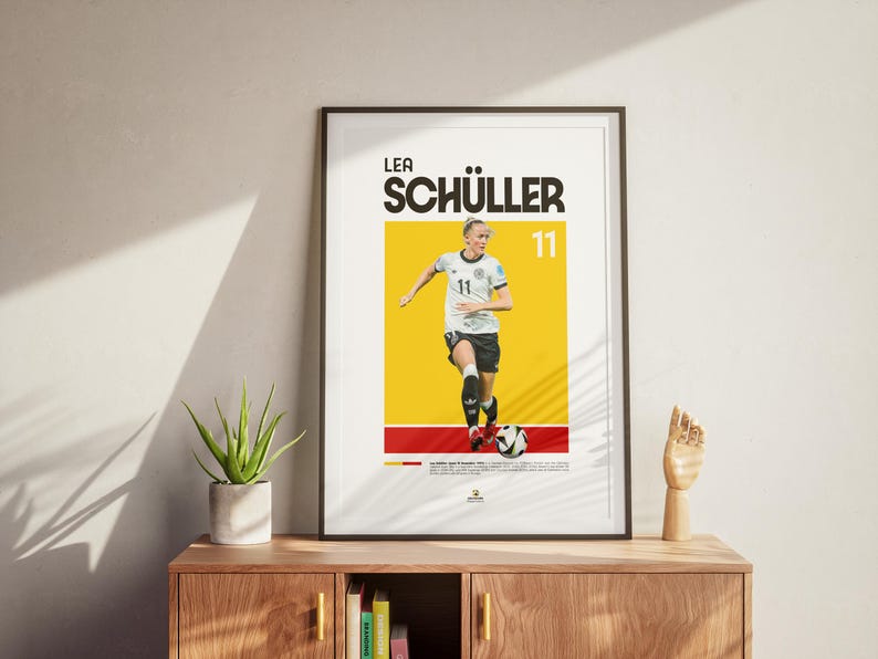 May include: Framed poster of footballer Lea Sch&uuml;ller in action, set against a yellow and red background. The poster displays her name and the number 11. The image is displayed on a neutral wall, with a wooden cabinet and plant in the foreground.