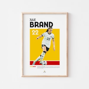 Może przedstawiać: Oprawiony plakat z piłkarką w biało-czarnym stroju, imieniem "JULE BRAND" i numerem "22" pogrubioną czcionką. Tło jest ż&oacute;łte i czerwone, z zawodniczką kopiącą piłkę nożną.
