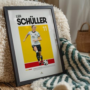 May include: Framed poster of Lea Sch&uuml;ller in a football uniform, number 11, set against a yellow and red backdrop. The black-framed poster rests on a textured surface, with a green and white blanket visible.