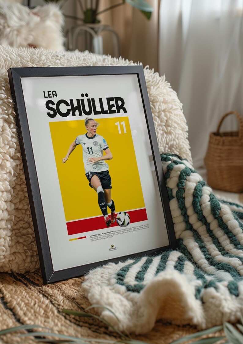 May include: Framed poster of Lea Sch&uuml;ller in a football uniform, number 11, set against a yellow and red backdrop. The black-framed poster rests on a textured surface, with a green and white blanket visible.