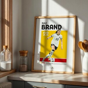 Może przedstawiać: Oprawiony w drewnianą ramę plakat z piłkarką w białej koszulce z numerem 22, na ż&oacute;łto-czerwonym tle. Plakat zawiera imię i nazwisko "JULE BRAND" czarnymi literami. Obraz jest w drewnianej ramie.