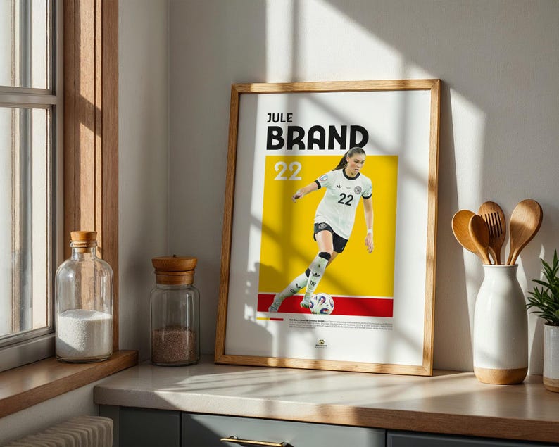 Może przedstawiać: Oprawiony w drewnianą ramę plakat z piłkarką w białej koszulce z numerem 22, na ż&oacute;łto-czerwonym tle. Plakat zawiera imię i nazwisko "JULE BRAND" czarnymi literami. Obraz jest w drewnianej ramie.