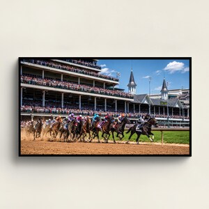May include: A framed print of a horse race in progress. Multiple horses with jockeys in colorful silks race on a dirt track. A large crowd fills the grandstand under a blue sky.