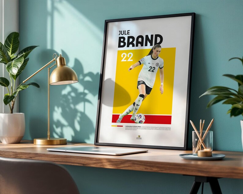 Może przedstawiać: Oprawiony plakat z piłkarką Jule Brand, numer 22, w biało-czarnym stroju. Plakat ma ż&oacute;łto-czerwone tło z nazwiskiem zawodnika pogrubionymi czarnymi literami. Plakat jest eksponowany na drewnianym biurku.