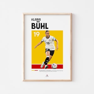 May include: Framed poster featuring Klara Bühl, a soccer player, in a white and black uniform with the number 19. The poster has a yellow and red background with the player in action, kicking a soccer ball. The player's name is at the top.