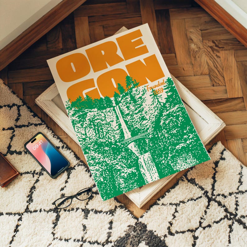 May include: A framed poster with the word "OREGON" in large orange letters. The poster features a green and white illustration of a forest and waterfall. The text "Founded 1859" is also visible. A smartphone, glasses, and a wallet are nearby.