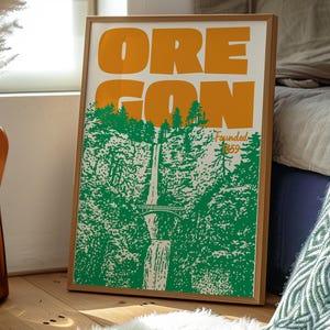 May include: Framed poster featuring the word "OREGON" in large orange letters above a green and white illustration of a waterfall and forest. The text "Founded 1859" is also present. The poster is in a wooden frame.
