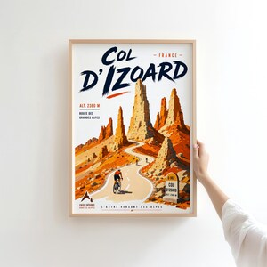 May include: A framed vertical poster featuring a stylised illustration of the Col d'Izoard in France. The artwork depicts a winding road through rocky orange peaks with a cyclist, a road sign reading D 902 Col d'Izoard, and text including Alt. 2360 M.