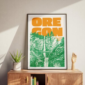 May include: Framed art print featuring the word "OREGON" in orange block letters above a green and white illustration of a waterfall and forest. The print is in a black frame and rests on a wooden cabinet with a plant and a wooden hand sculpture.