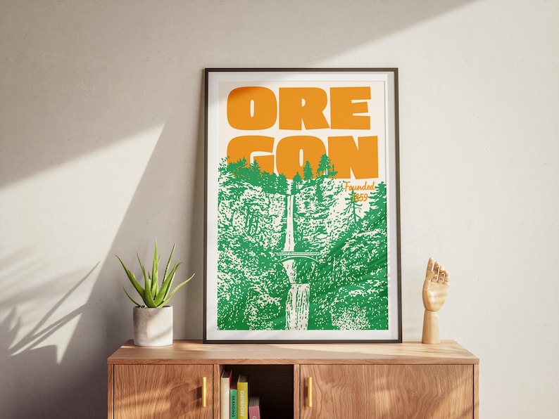 May include: Framed art print featuring the word "OREGON" in orange block letters above a green and white illustration of a waterfall and forest. The print is in a black frame and rests on a wooden cabinet with a plant and a wooden hand sculpture.