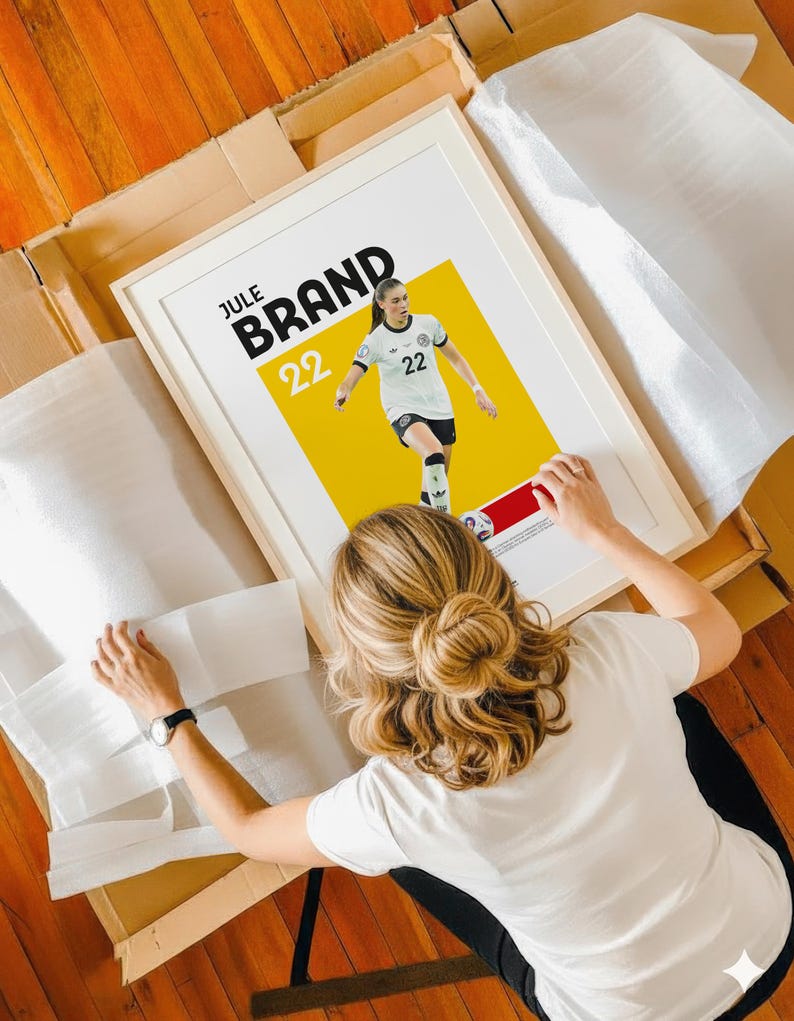 Może przedstawiać: Oprawiony plakat z piłkarką w biało-czarnym stroju, z napisem "JULE BRAND" i numerem "22" pogrubioną czcionką. Tło jest ż&oacute;łte i czerwone. Plakat jest w trakcie obsługi.