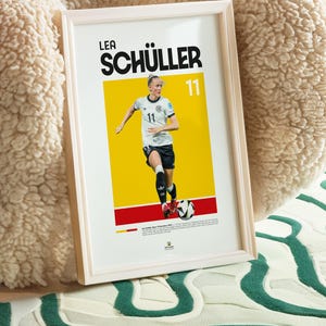 May include: Framed poster of footballer Lea Sch&uuml;ller in a white and black kit, number 11. The background is yellow and red. The poster is in a white frame, ideal for a sports fan's decor.