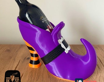 Puede incluir: Porta botellas de vino con forma de zapato de bruja morado con un tacón rayado en negro y naranja. El porta botellas está lleno con una botella de vino tinto.