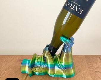 May include: A green, blue, and gold 3D printed wine bottle holder shaped like a person holding a bottle of wine. The bottle is labeled "BATON" Marlborough Sauvignon.