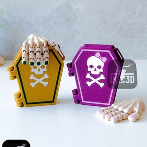 May include: Two coffin-shaped boxes, one yellow and one purple, with skull and crossbones designs. The yellow box has a skeleton hand on top. The purple box has a skull with a bow. A detached skeleton hand is also present.