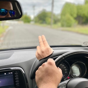 May include: A person driving a car with their hand raised in the air. The car has a black steering wheel with a Mazda logo. The dashboard shows a digital clock displaying 1:45.
