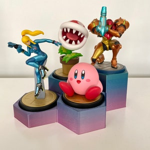 Può includere: Un set di quattro figurine di personaggi di videogiochi esposte su un supporto esagonale. Il supporto è realizzato in plastica con un gradiente di colore viola e blu. Le figurine includono un Kirby rosa, una pianta Piranha verde, un Samus Aran dorato e un Zero Suit Samus blu e giallo.