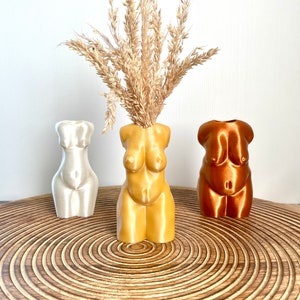 Può includere: Tre vasi in ceramica a forma di torso umano, in bianco, giallo e rame. Il vaso giallo contiene erba della pampa secca.