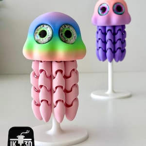 May include: Two whimsical 3D-printed jellyfish sculptures. One features a rainbow-colored head and pink body, the other has a pink and purple head and a purple body. Both have large, detailed eyes and are mounted on white stands.