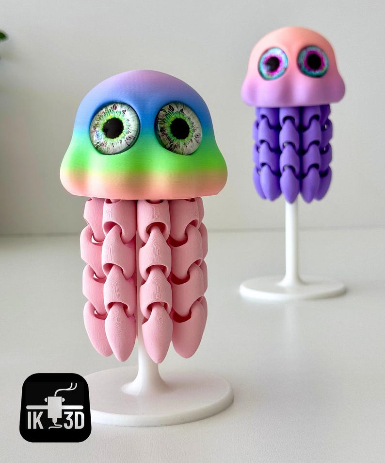 May include: Two whimsical 3D-printed jellyfish sculptures. One features a rainbow-colored head and pink body, the other has a pink and purple head and a purple body. Both have large, detailed eyes and are mounted on white stands.