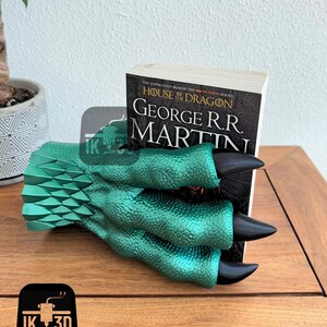 May include: A green dragon claw bookend holding a book titled "House of the Dragon" by George R.R. Martin. The bookend is made of a textured, metallic green material with black claws.