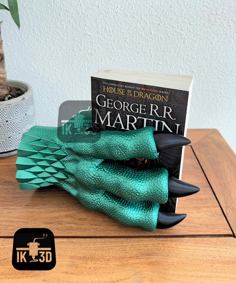May include: A green dragon claw bookend holding a book titled "House of the Dragon" by George R.R. Martin. The bookend is made of a textured, metallic green material with black claws.