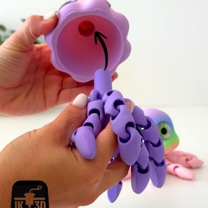 May include: A 3D-printed articulated octopus toy in shades of purple, pink, and rainbow colors. The octopus has a bell-shaped head and jointed tentacles. The head is held open, revealing a red ball inside. The logo "IK 3D" is in the bottom left corner.