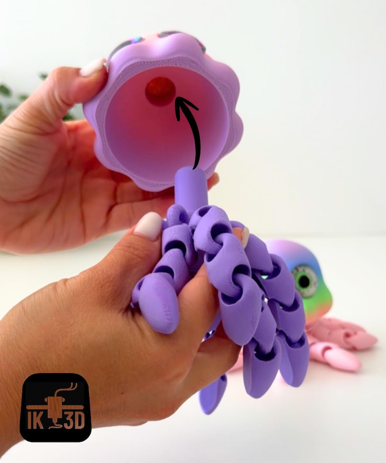 May include: A 3D-printed articulated octopus toy in shades of purple, pink, and rainbow colors. The octopus has a bell-shaped head and jointed tentacles. The head is held open, revealing a red ball inside. The logo "IK 3D" is in the bottom left corner.