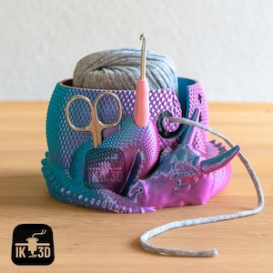 Puede incluir: Un cuenco de hilo impreso en 3D de color morado y turquesa con forma de rinoceronte con una bola de hilo gris, un par de tijeras y un gancho de ganchillo.
