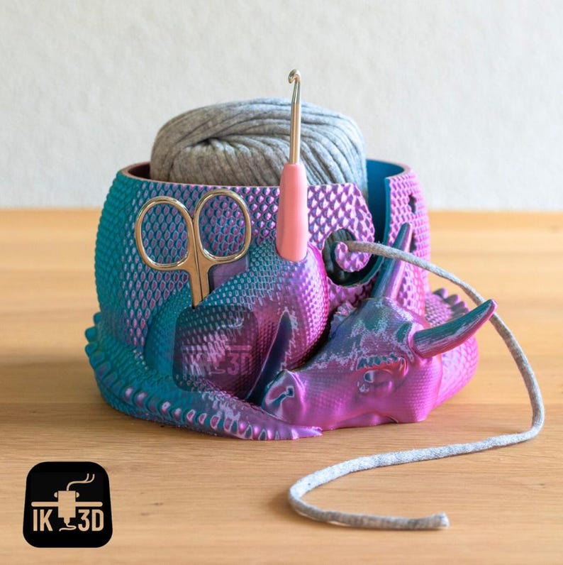 May include: A purple and teal 3D printed yarn bowl shaped like a rhinoceros with a grey yarn ball, a pair of scissors, and a crochet hook.