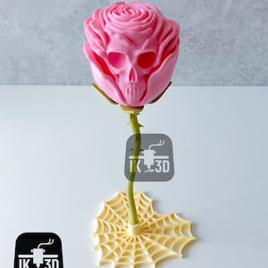 Op de afbeelding: Een roze roos sculptuur met een schedel ontwerp in het midden van de bloem. De stengel is groen en de basis is een geel spinnenweb ontwerp. Het logo "IK 3D" is zichtbaar.