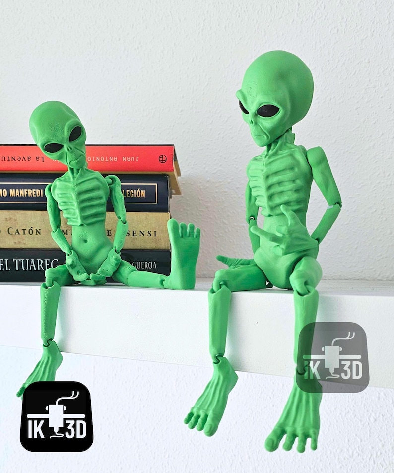Puede incluir: Dos figuritas de extraterrestres verdes con extremidades articuladas, sentadas en un estante blanco. Los extraterrestres tienen cabezas grandes con ojos negros y una apariencia esquel&eacute;tica.