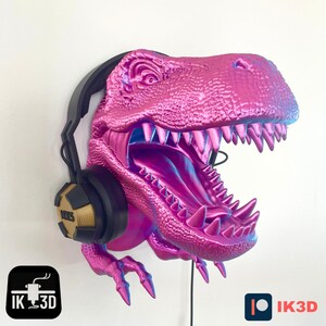 May include: A pink dinosaur head sculpture with a black and gold headphone attached. The dinosaur has a large open mouth with sharp teeth and a textured surface. The sculpture is mounted on a wall.