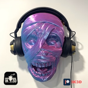 May include: A 3D printed mummy head wearing black headphones with gold accents. The mummy head is a light blue and pink colour with a glittery finish. The headphones are connected to a black cord.