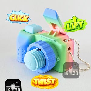 Puede incluir: Un llavero de cámara impreso en 3D de colores azul, verde y rosa. La cámara tiene una lente, una parte superior que se levanta y las palabras "CLICK", "LIFT" y "TWIST" en bocadillos. Se adjunta una cadena plateada.