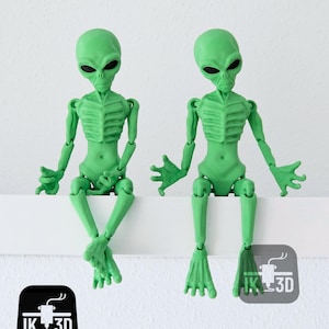 Puede incluir: Dos figuras de extraterrestres verdes con extremidades articuladas, sentadas sobre una superficie blanca. Los extraterrestres tienen cabezas grandes, ojos negros y estructuras esquel&eacute;ticas detalladas. Las figuras est&aacute;n impresas en 3D y tienen un acabado mate.