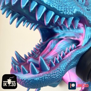 May include: Close-up of a 3D printed dinosaur head with a blue and pink iridescent finish. The dinosaur's mouth is open, revealing sharp teeth and a pink tongue.